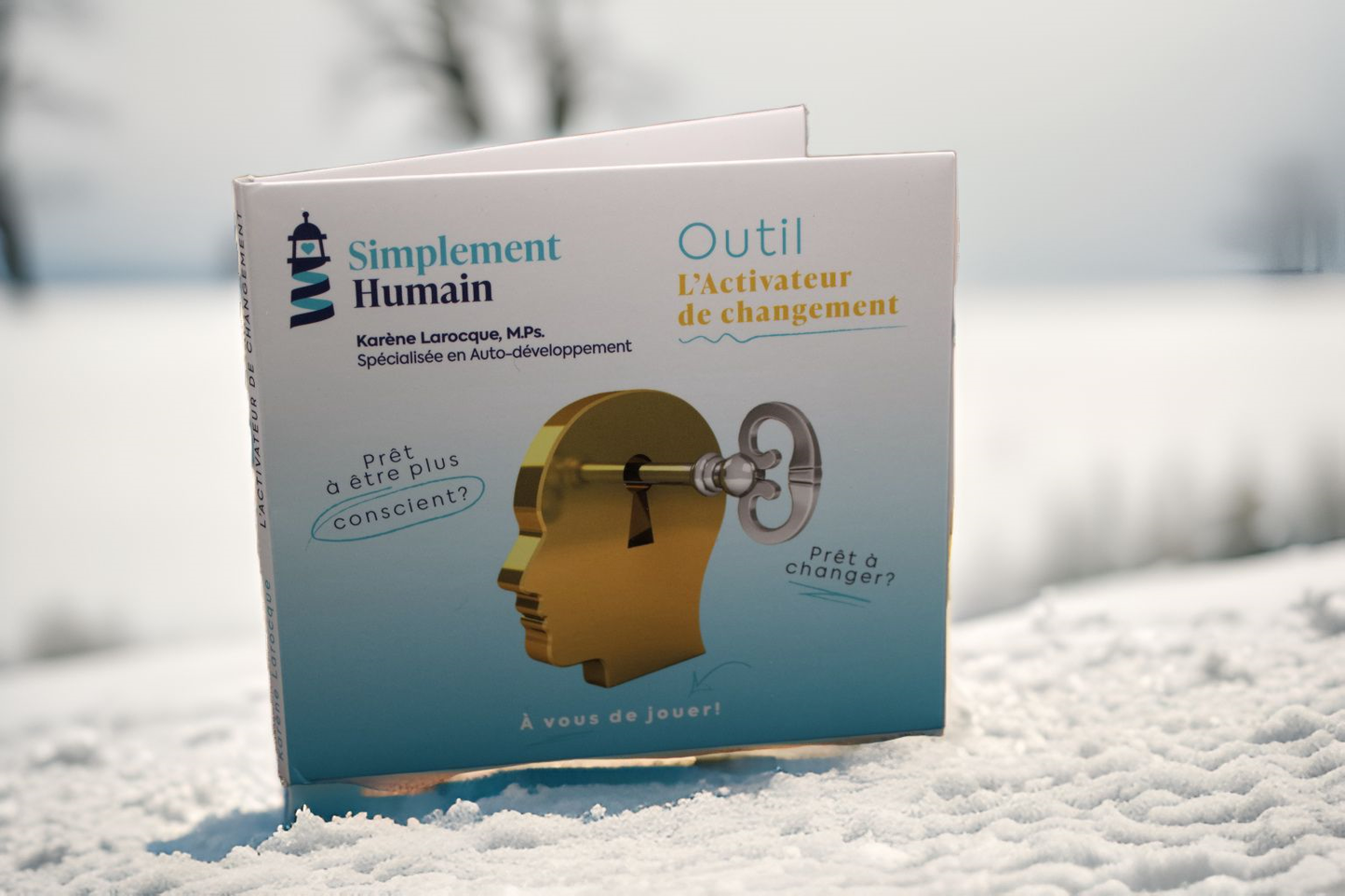 Photo de l'outil "L'Activateur de changement" de Simplement Humain, présenté dans un décor hivernal. Le visuel montre un design de tête dorée avec une clé, accompagné des phrases : "Prêt à être plus conscient ?" et "Prêt à changer ?" sur un fond blanc.