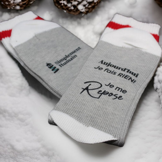 Chaussettes grises Simplement Humain avec le message 'Aujourd'hui je fais rien, je me repose', posées dans la neige avec des éléments décoratifs d'hiver.
