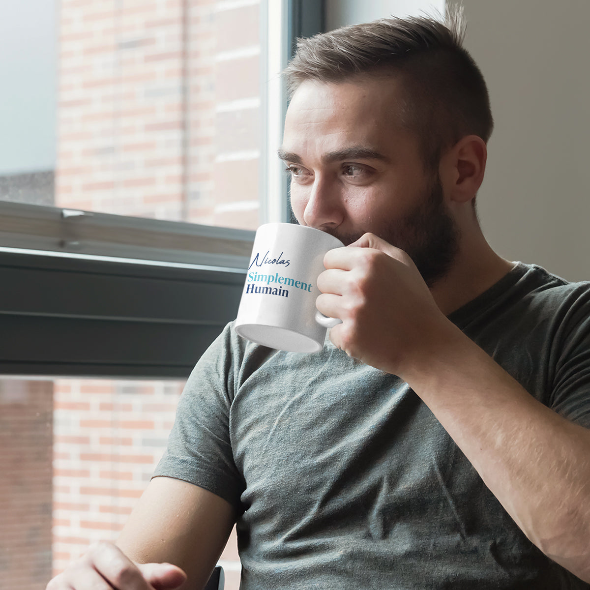 Homme souriant buvant dans une tasse blanche personnalisée avec le prénom "Nicolas" et le logo Simplement Humain, assis près d'une fenêtre.