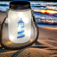 Lanterne lumineuse en verre avec le logo Simplement Humain, éclairée de petites lumières LED, posée sur le sable au bord de la plage au coucher du soleil.