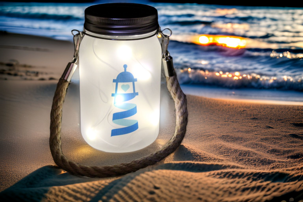 Lanterne lumineuse en verre avec le logo Simplement Humain, éclairée de petites lumières LED, posée sur le sable au bord de la plage au coucher du soleil.