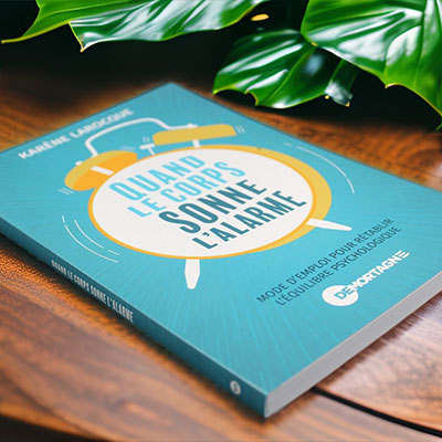 Photo du livre "Quand le corps sonne l'alarme" de Karène Larocque, posé sur une table en bois avec un feuillage vert en arrière-plan. Le livre se distingue par sa couverture turquoise et son design attrayant.